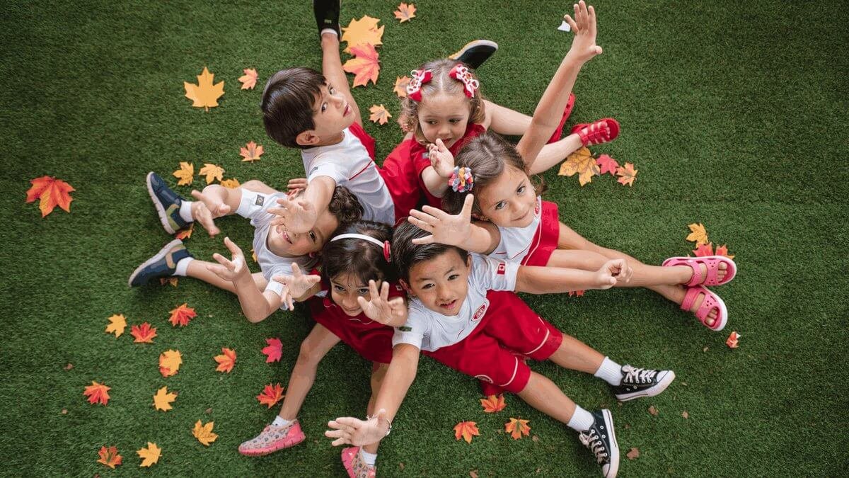 Vista superior de alunos da educação infantil sentados na grama, sorrindo e com folhas de plátano espalhadas pelo chão, no jardim da Maple Bear.